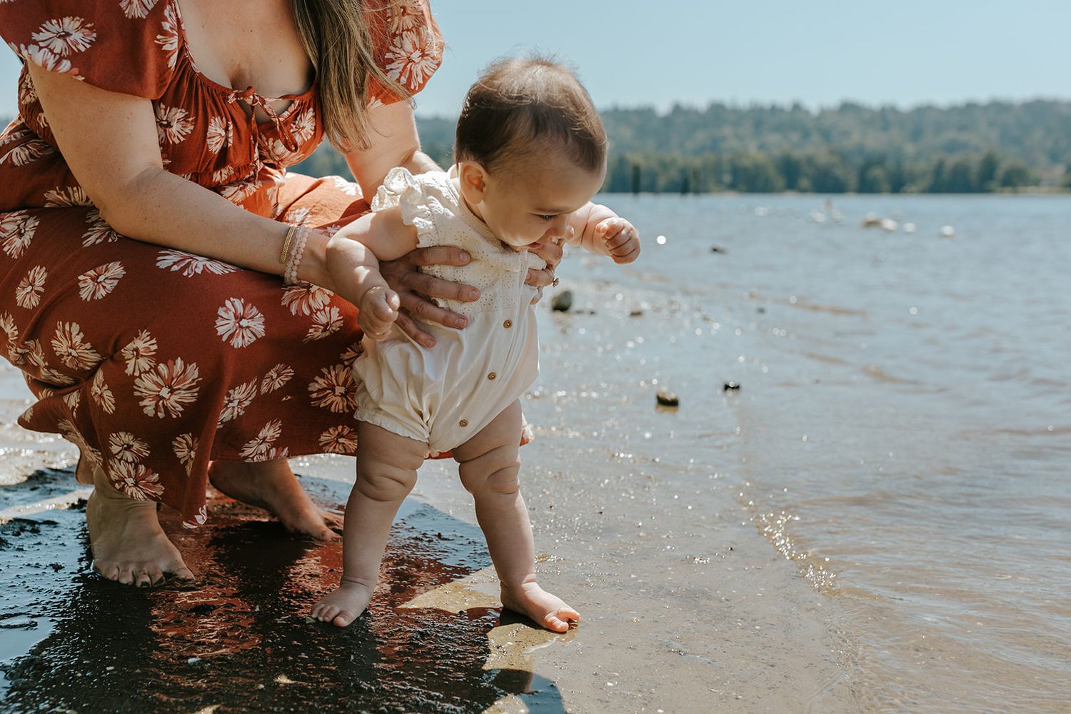 A baby takes first steps at the beach while being supported in shallow water.