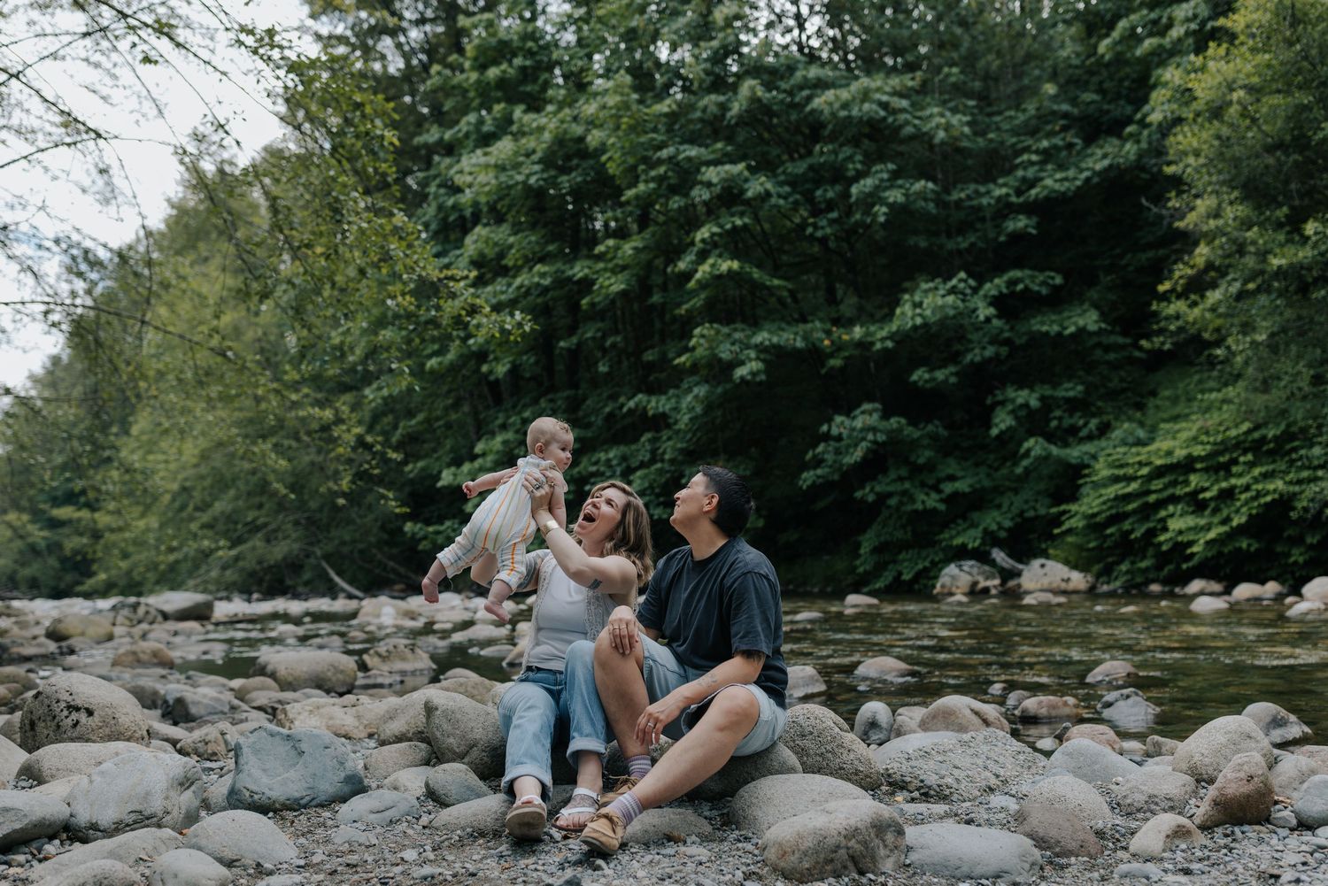 Family sitting on rocky riverbank with lush green forest backdrop, parents lifting baby joyfully outdoors.