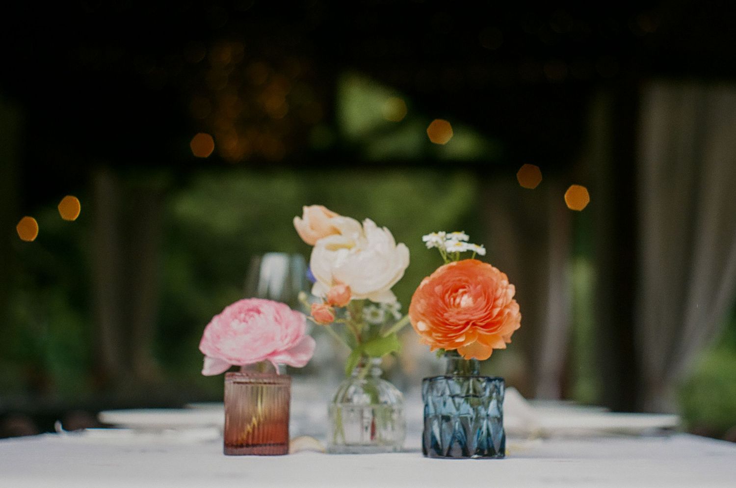 Delicate pink and orange flowers arranged in colorful glass vases on a white tablecloth.