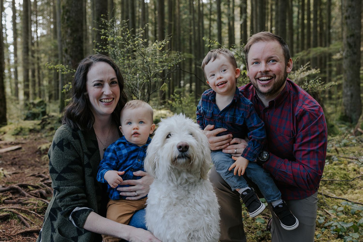 Family and their white fluffy dog pose together on a fallen log in a lush forest setting.