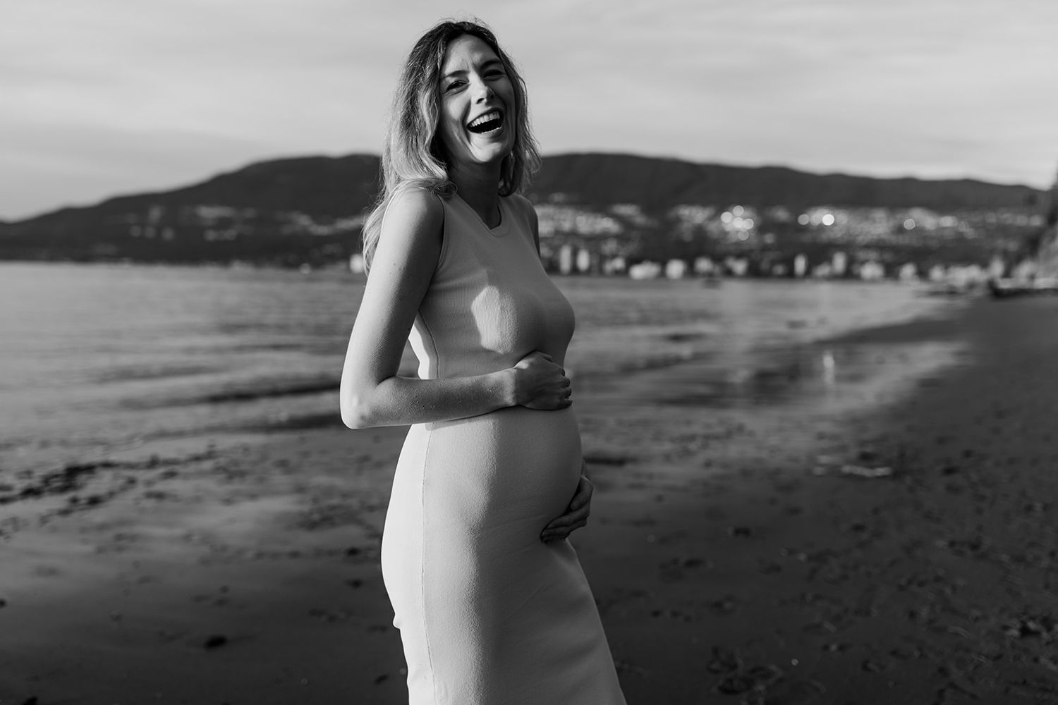 A silhouetted figure in a white dress stands on a beach during a maternity photoshoot.