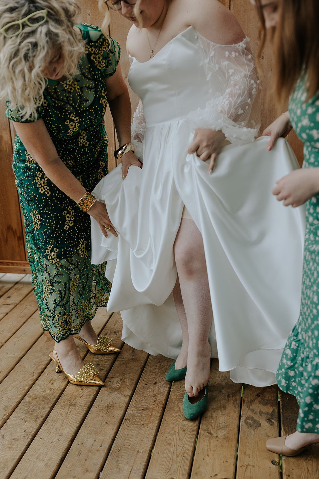 Wedding party members in green dresses helping with shoe fittings on wooden deck.