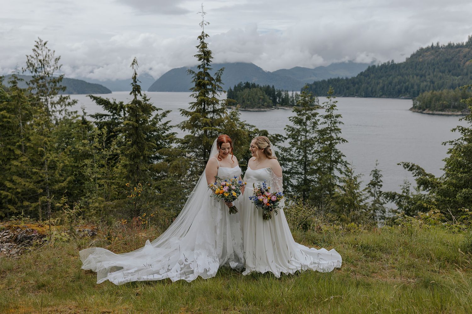 Two wedding figures stand against a dramatic mountain lake backdrop holding colorful bouquets.