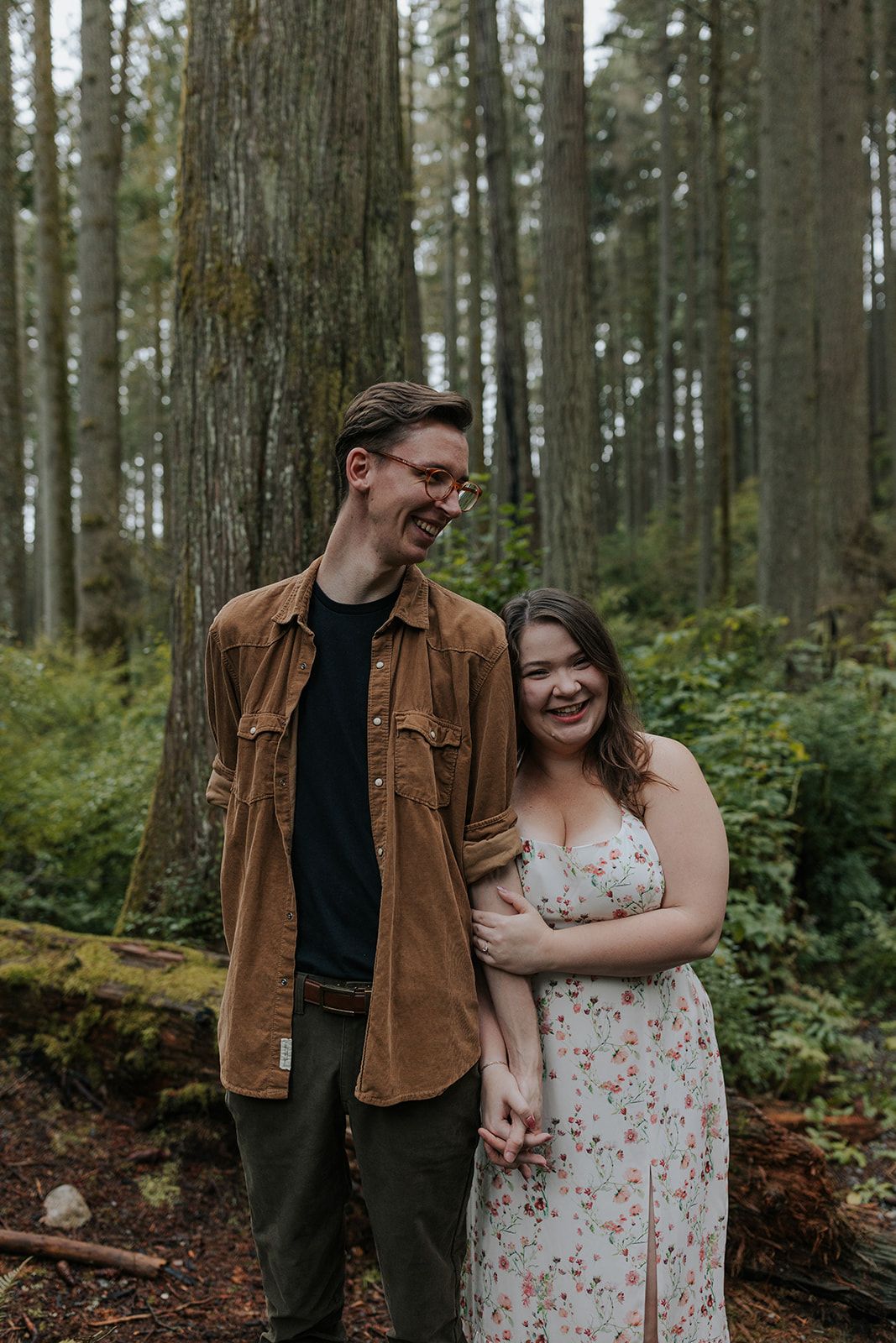 Couple laughing together during a casual photoshoot in a peaceful forest setting.