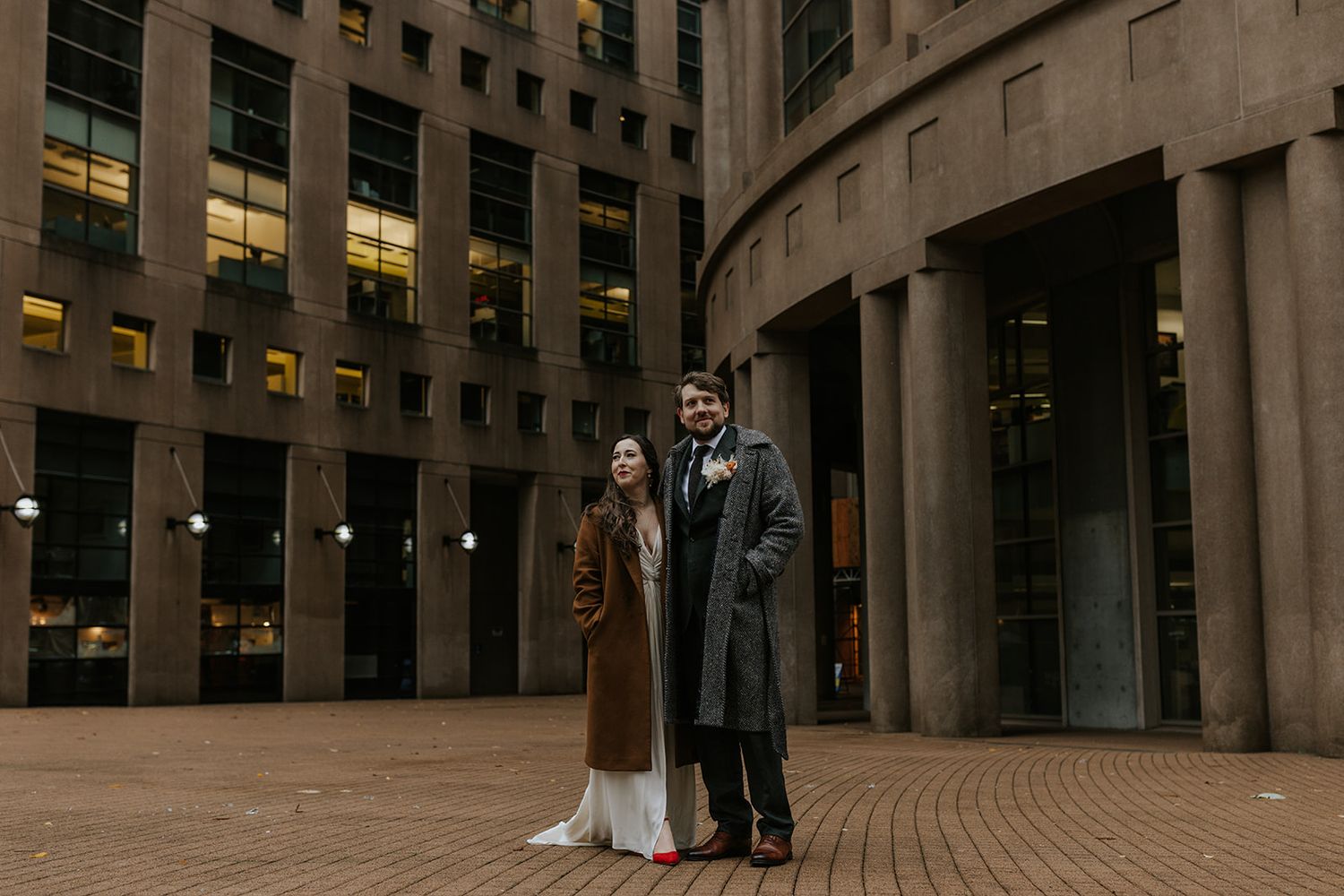 A couple stands together in formal winter coats outside a modern curved office building at dusk.