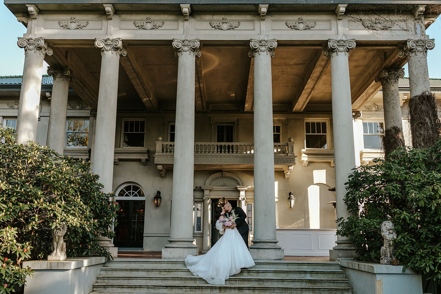 A bride in a white wedding gown stands in an elegant colonnade walkway with classical architectural details.