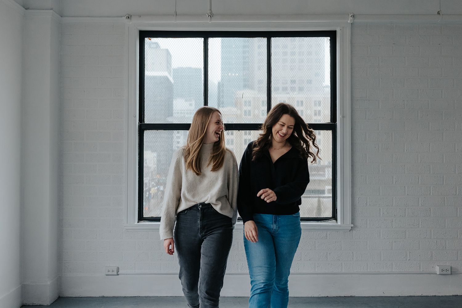 Two people walking and laughing in a bright white studio space with industrial windows.