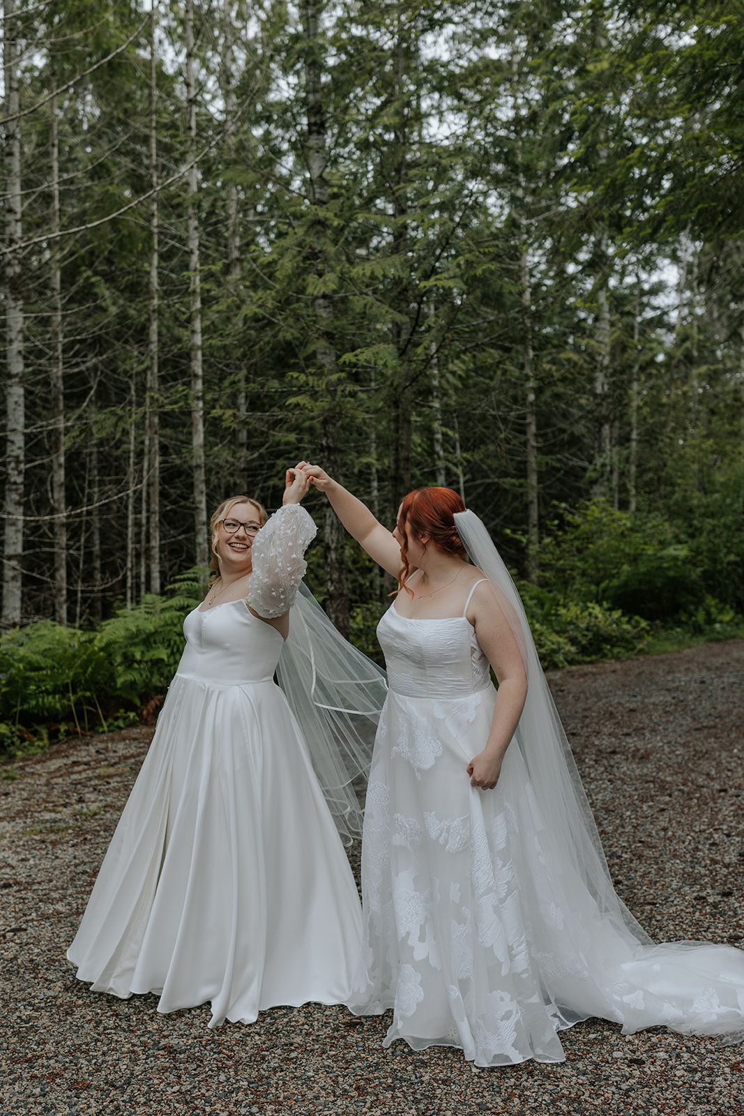 Two brides in flowing white gowns dance together on a forest path.