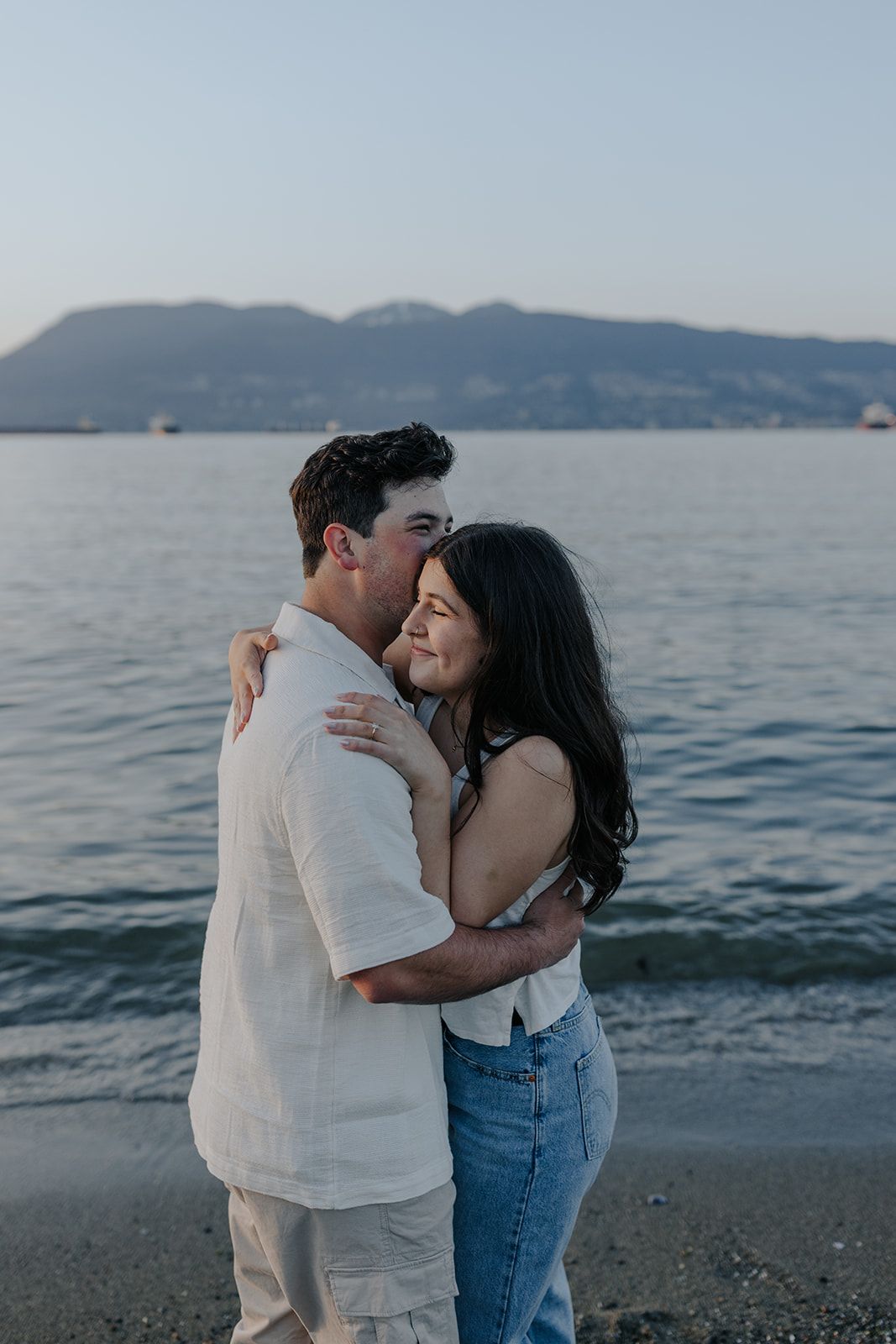 A couple embraces on a beach at dusk with mountains and water in the background.