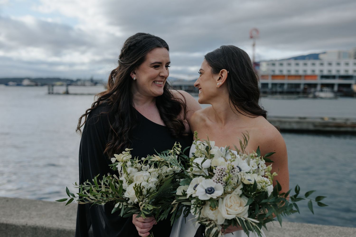 Wedding party poses along waterfront with white bouquets and formal attire on overcast day.