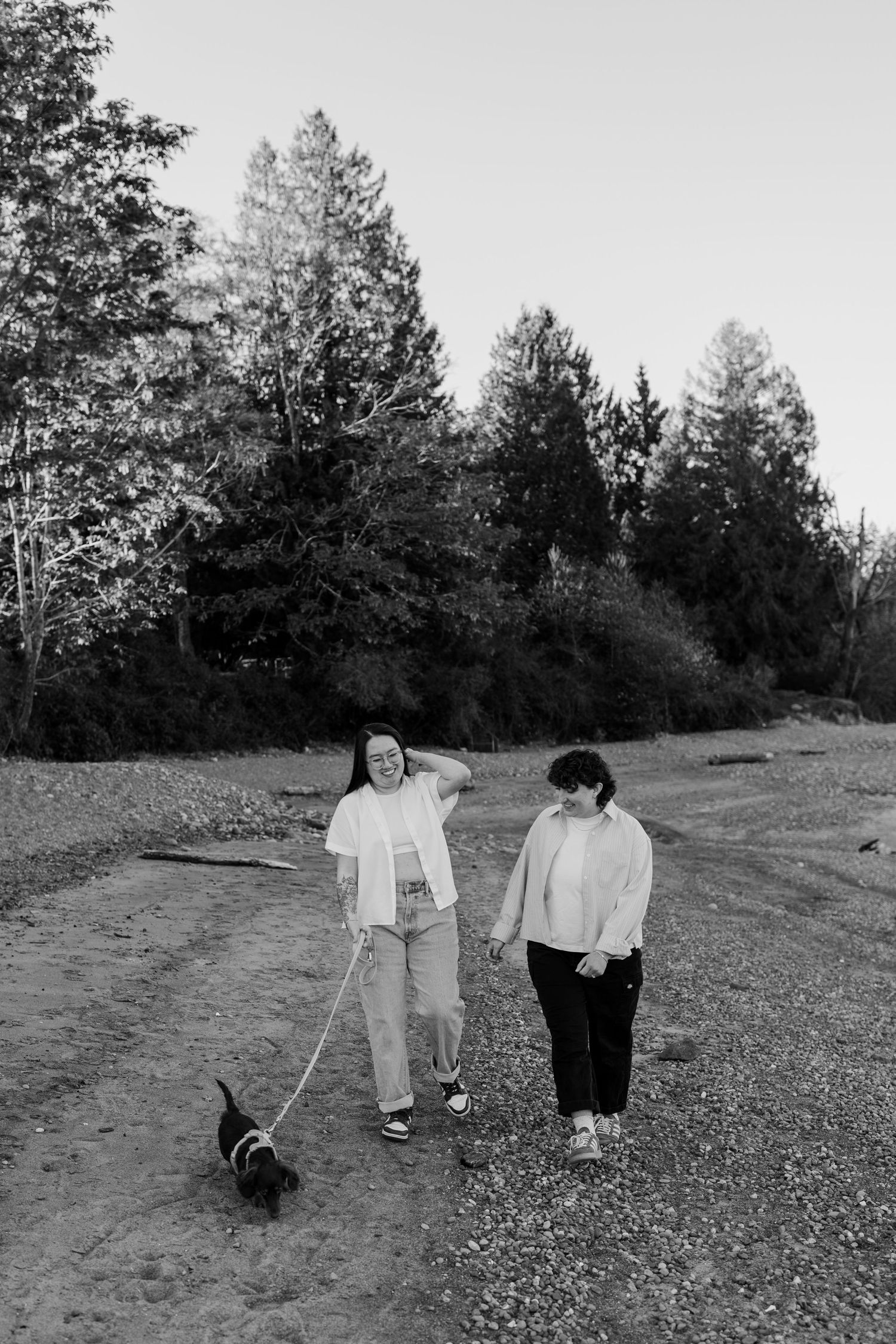 Two women walking a small dog on a leash along a gravel path surrounded by tall trees in black and white.