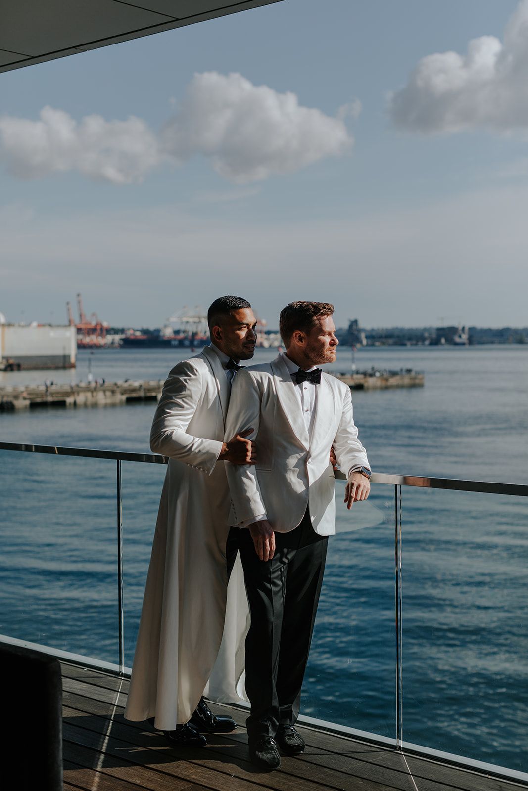 Two people in white jackets lean against a railing overlooking a harbor on a sunny day.