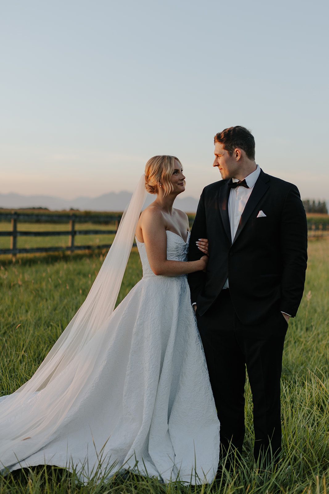 A bride in a flowing white gown and groom in black suit pose in an open field at sunset.
