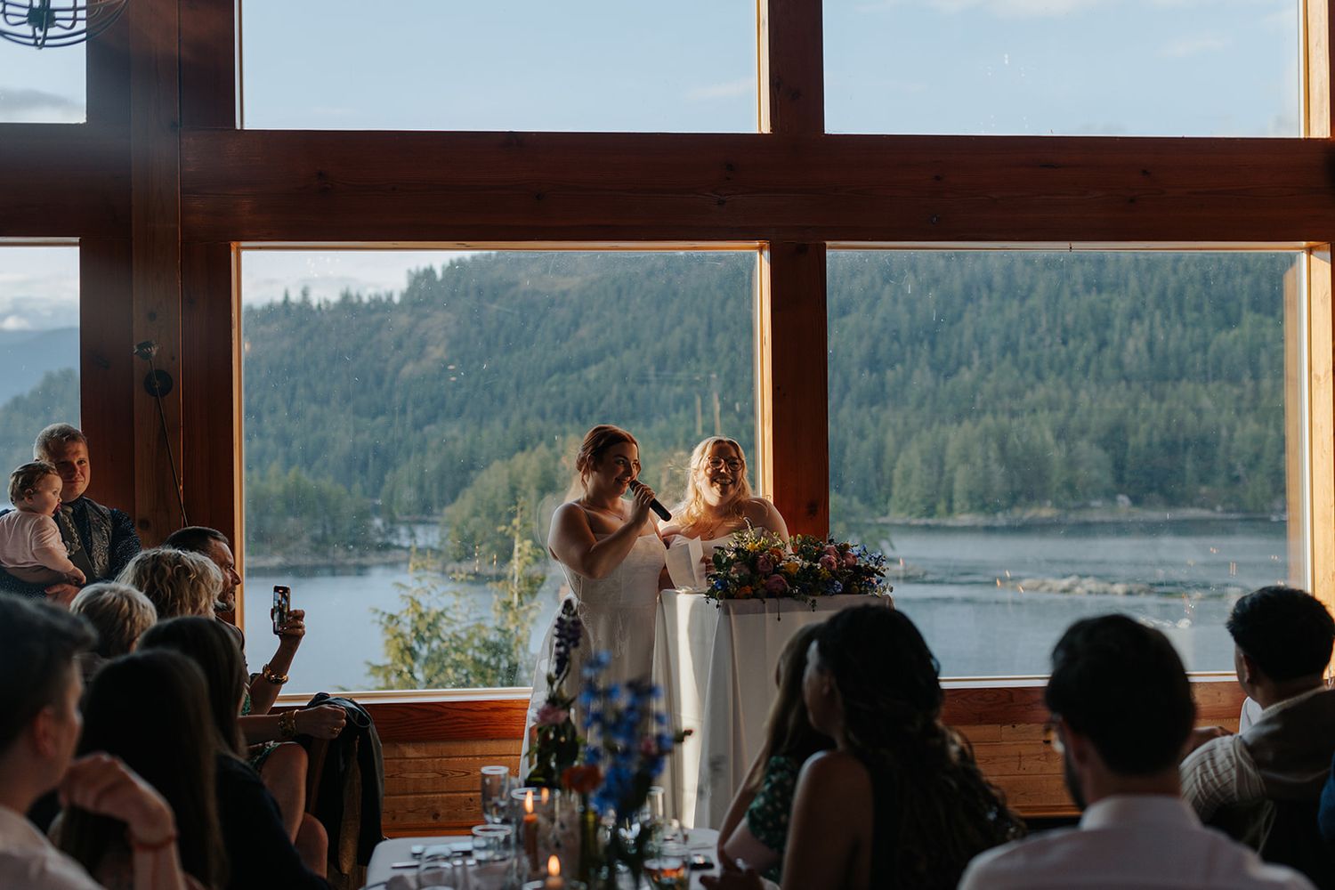 Silhouetted figures dance and celebrate inside a rustic waterfront venue with large windows overlooking a scenic lake view.