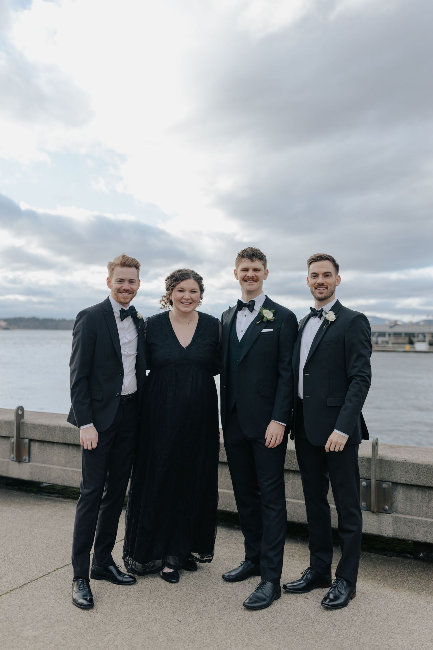 A wedding party in formal black attire poses together on a waterfront promenade.