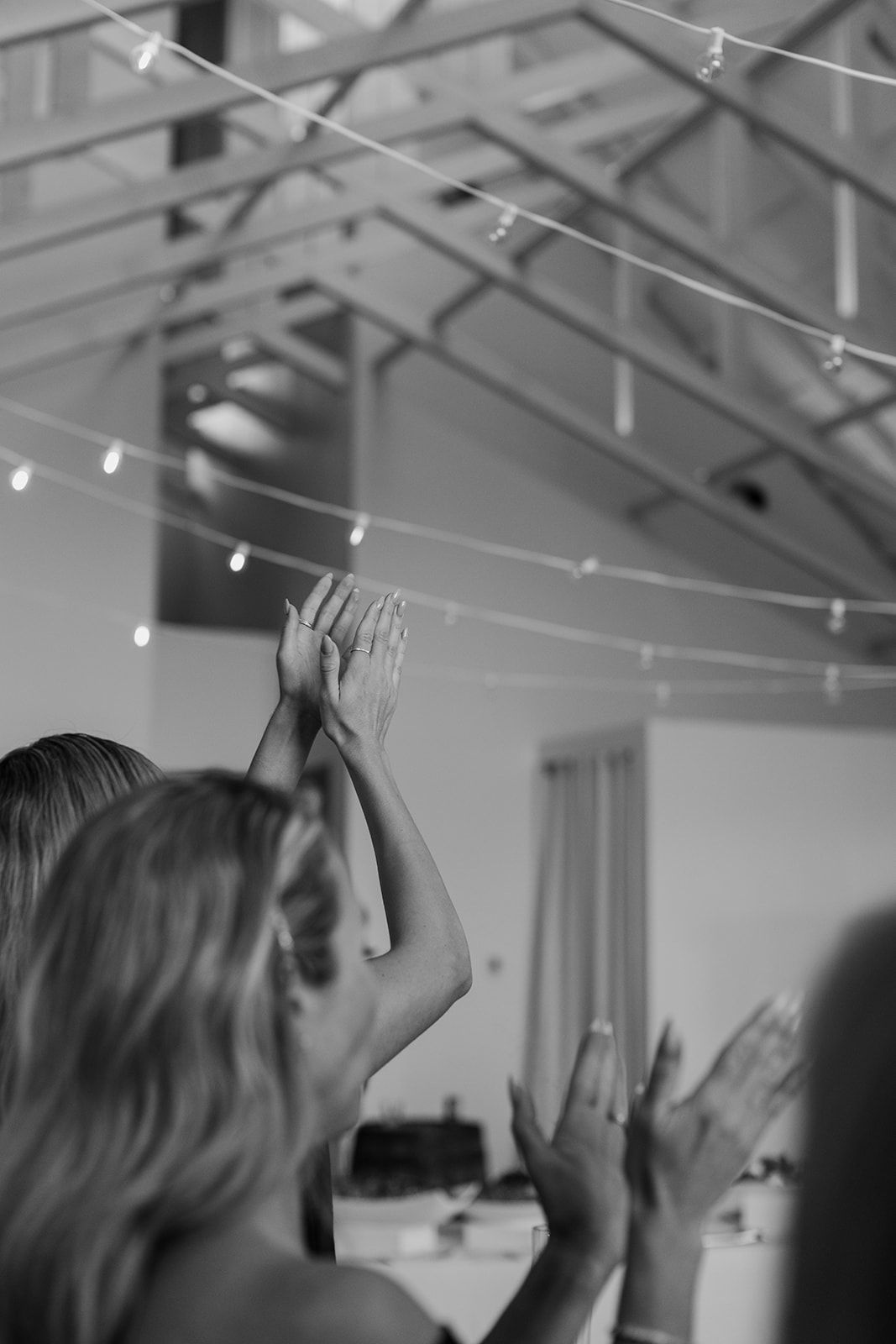 Wedding guests raise their hands in celebration under string lights at an indoor reception venue.