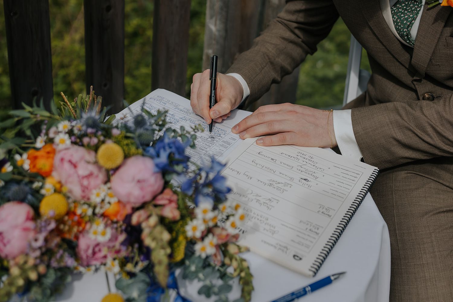 A guest signs the wedding guestbook beside a floral arrangement on a rustic table.