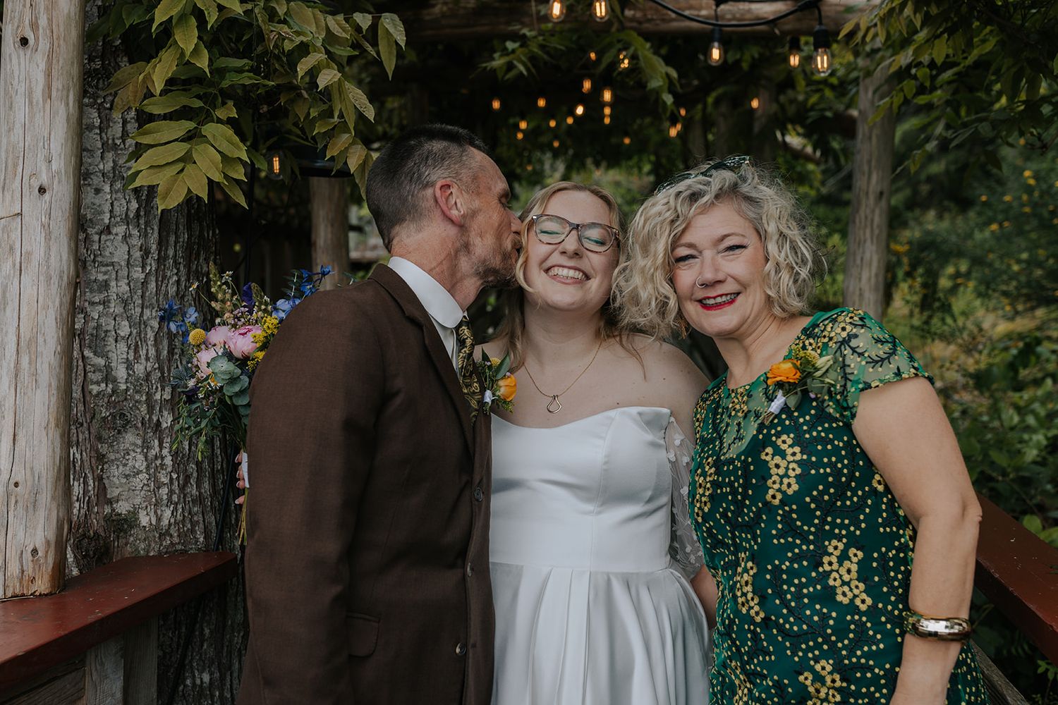 Three people share an intimate moment during wedding celebration under garden pergola.
