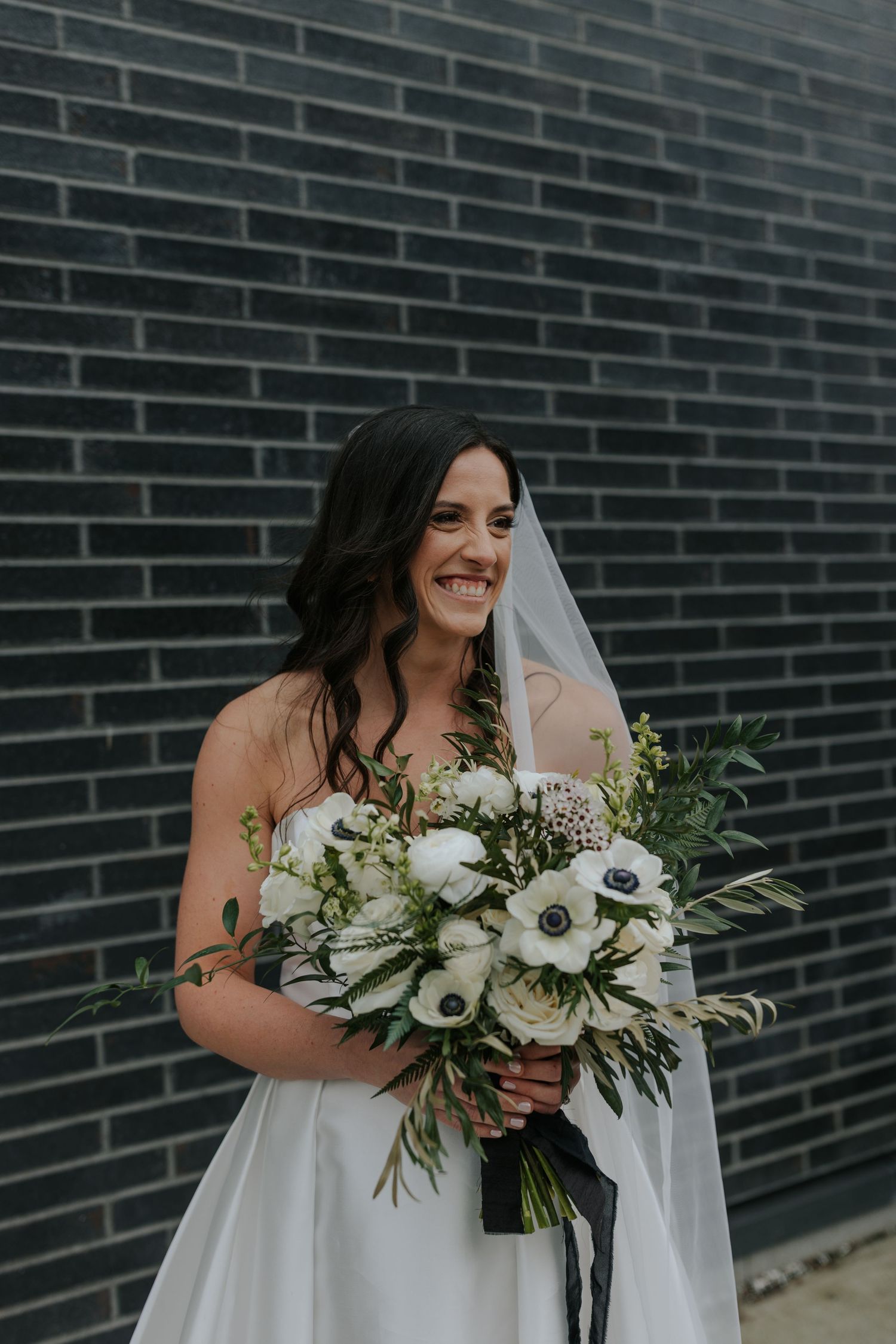 Bride in white dress holds stunning white and green bouquet against modern brick wall.