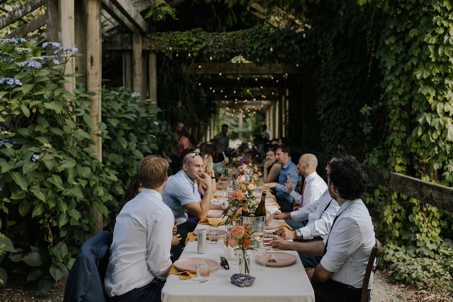 A group of people enjoying an outdoor dining experience at a long table surrounded by lush greenery.