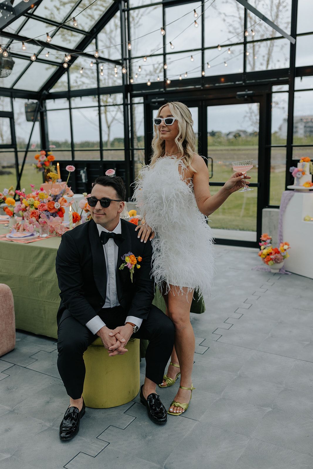 A couple in sunglasses and formal attire pose playfully in a modern glass greenhouse with colorful floral decorations.