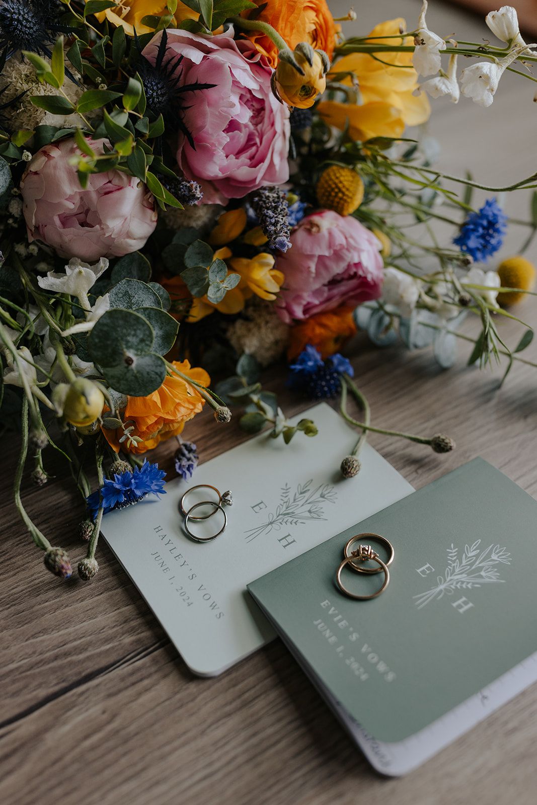 Colorful wedding flowers with rings and invitation cards on wooden surface.