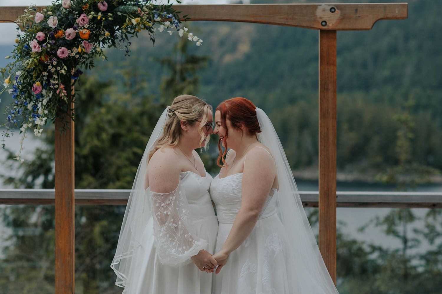Two brides in white wedding dresses share an intimate moment under a wooden arch with mountain scenery.