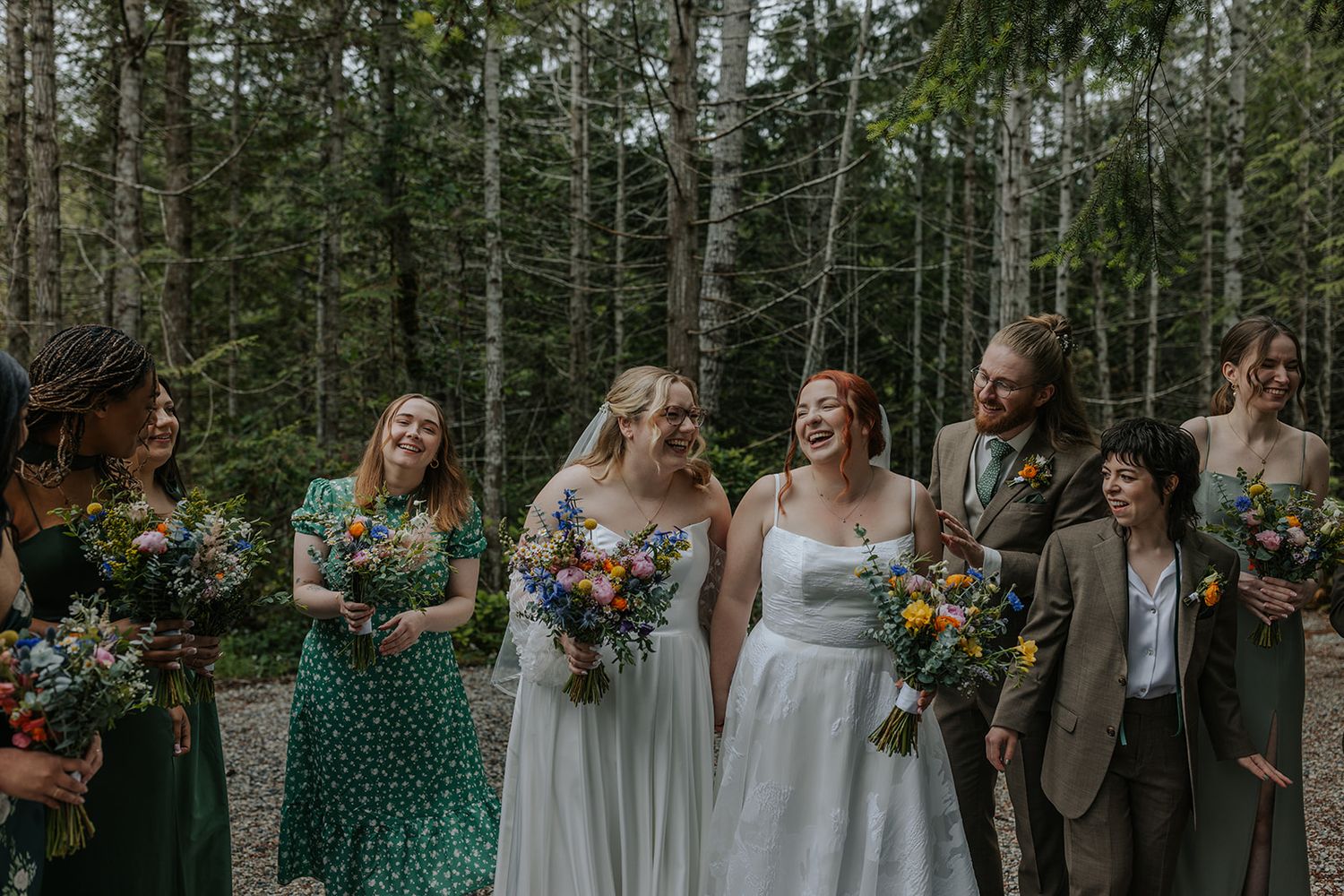 Bridal party in green and white dresses poses together in forest setting.