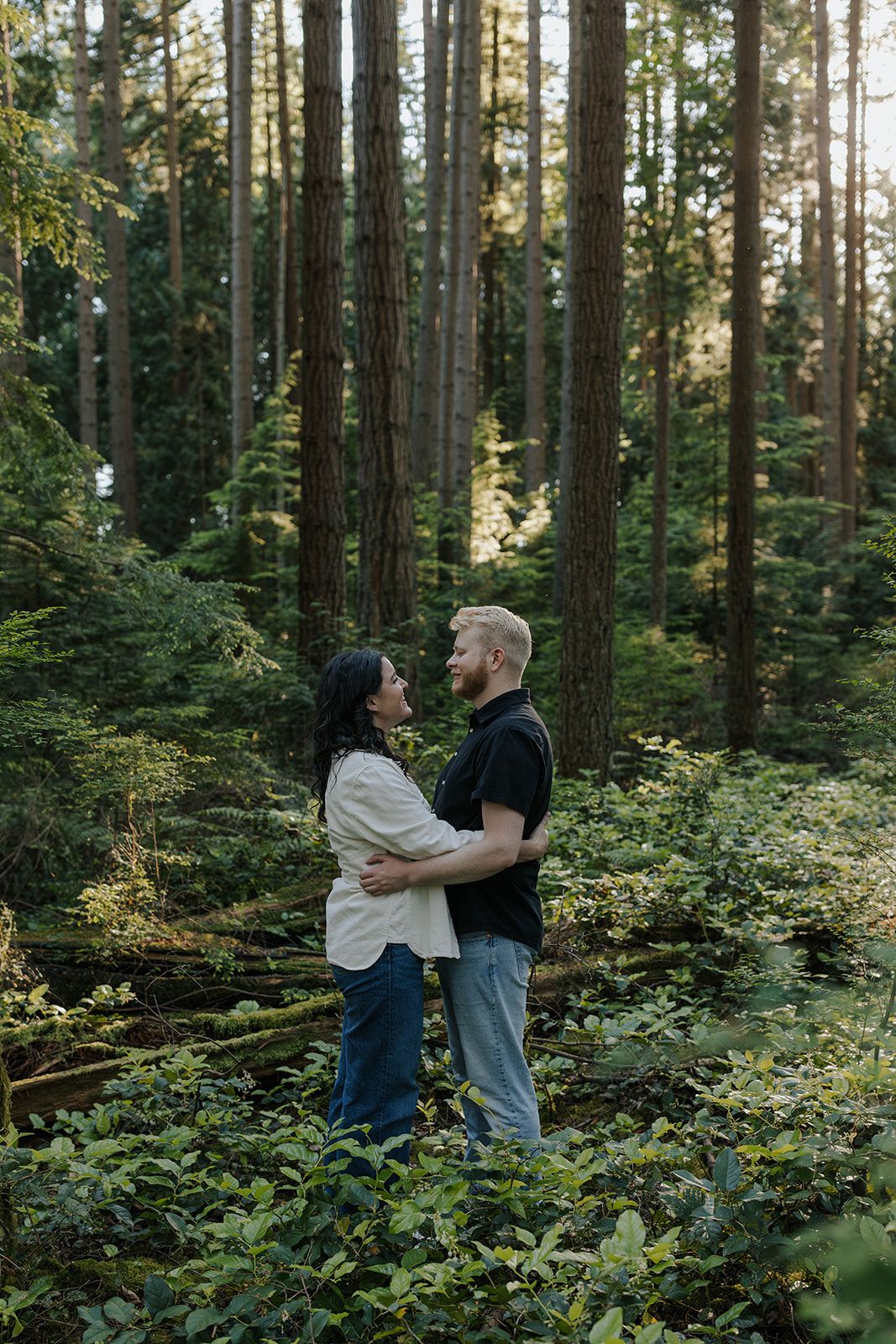 A couple embraces in a lush green forest with tall evergreen trees and dappled sunlight.