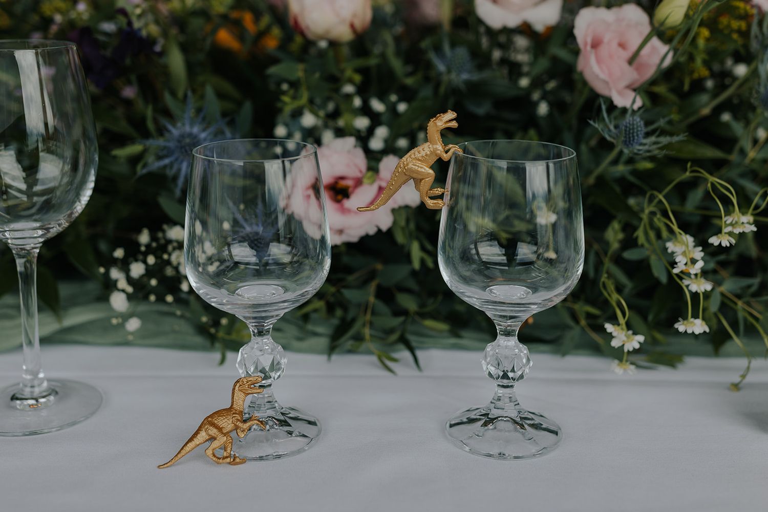 Wedding table setting with wine glasses and gold dinosaur decorations among floral arrangements.