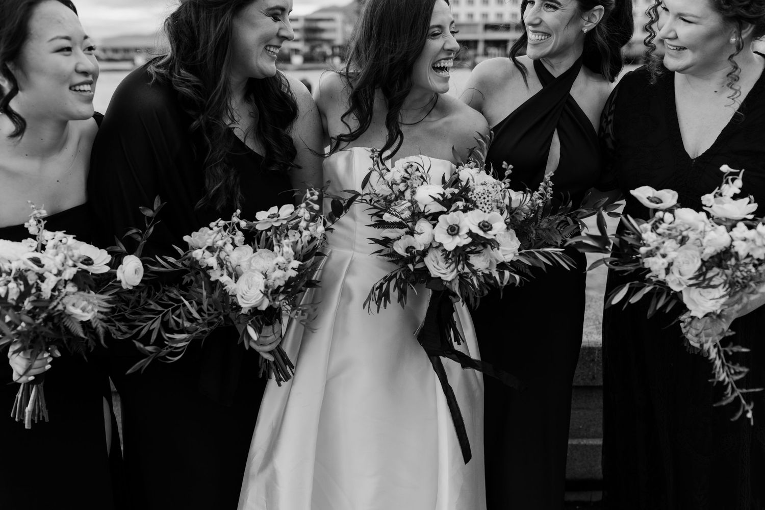 Wedding party in black dresses laughing together while holding floral bouquets.