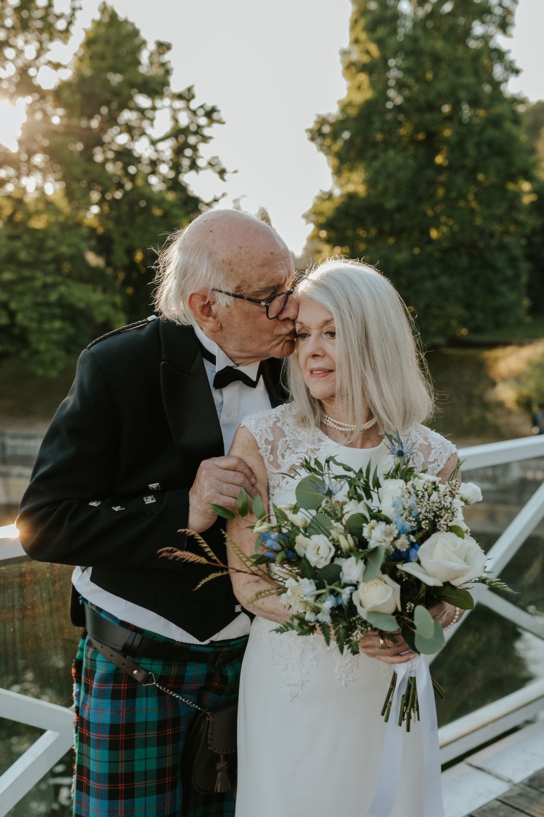 Romantic outdoor wedding moment with couple in white dress and tartan kilt at sunset.