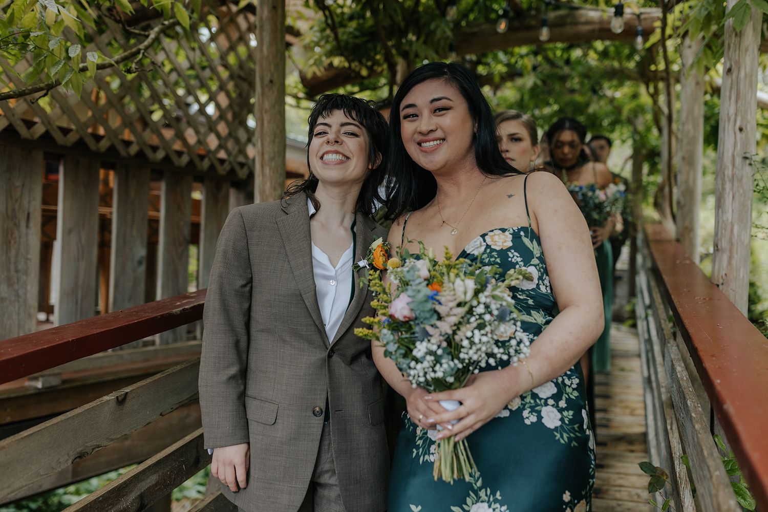 Two people share a joyful moment in a floral dress and suit on a wooden deck surrounded by greenery.