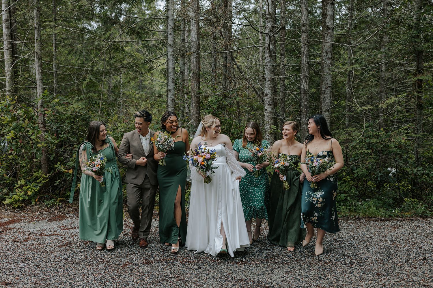 Wedding group poses casually on forest path surrounded by tall evergreen trees.