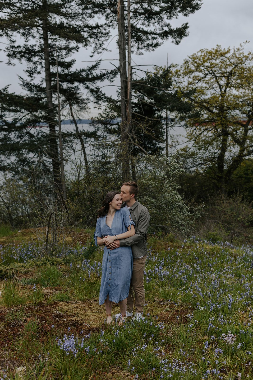 A couple stands together in a field of purple wildflowers with evergreen trees in the background.