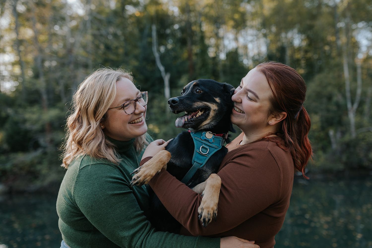 Two people holding and playing with a happy black dog wearing a blue bandana outdoors.