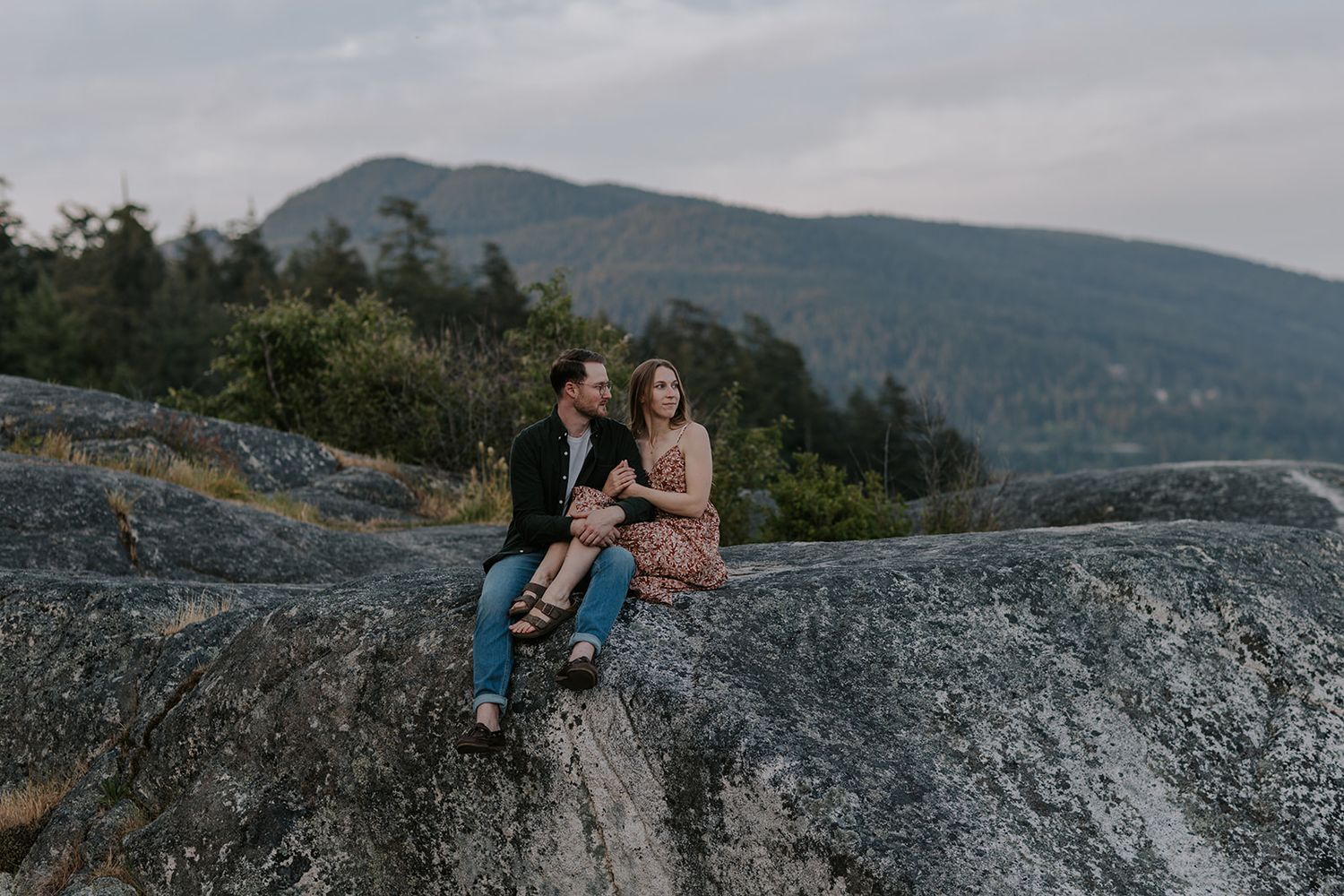 Couple sitting together on rocky mountain summit with lush green hills and cloudy sky in background.
