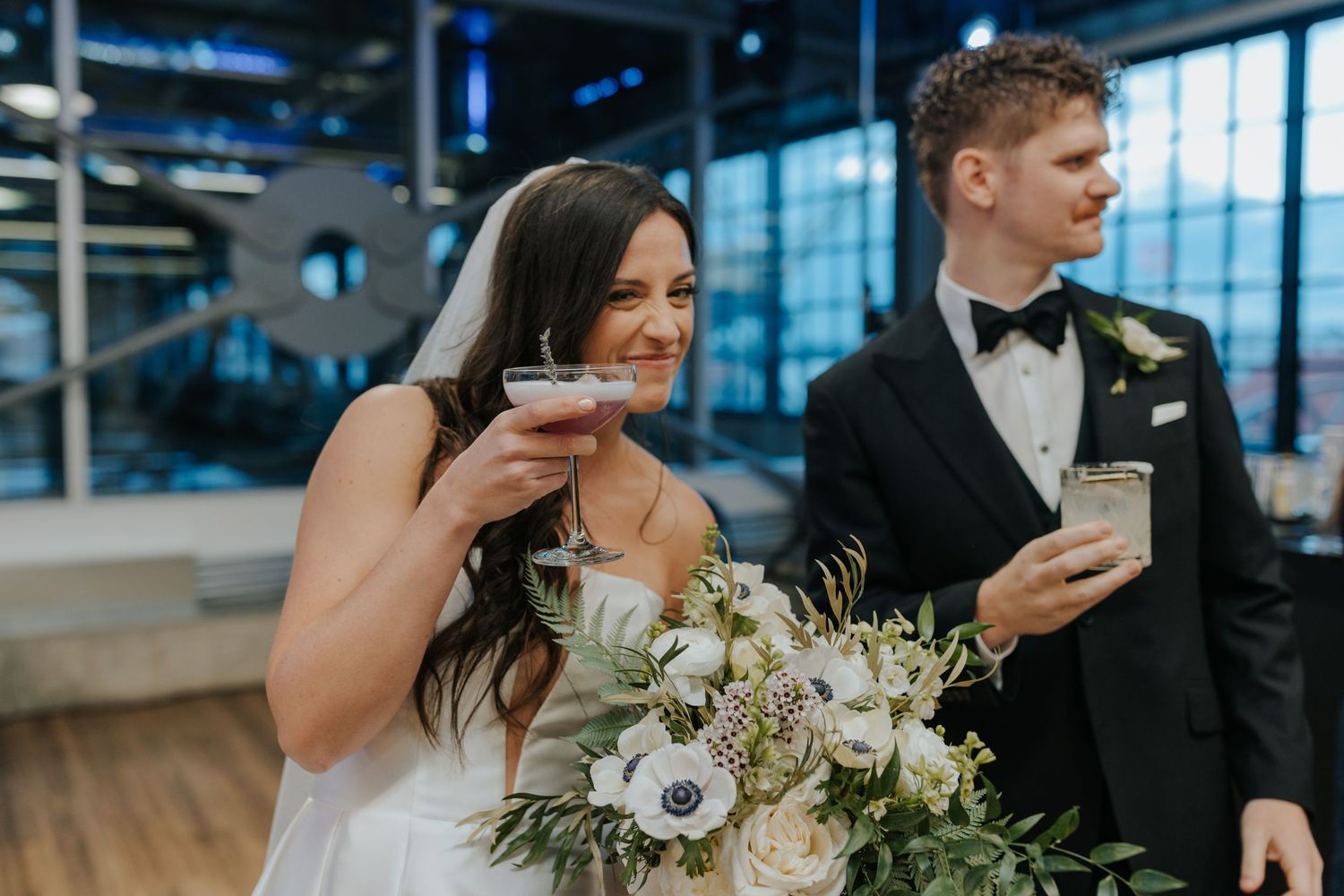 A couple enjoys cocktails together during their wedding reception in a modern venue with large windows.