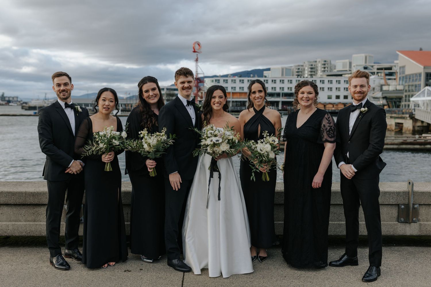 Wedding party in black and white attire gathered on pier with cloudy waterfront backdrop.