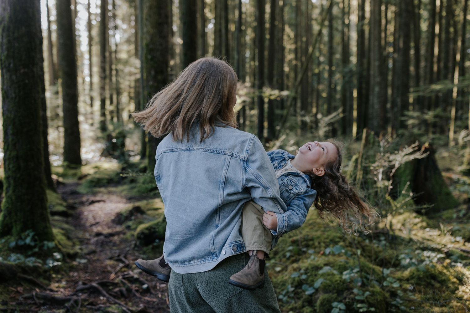 Woman carrying a laughing child through a lush green forest path surrounded by tall trees.