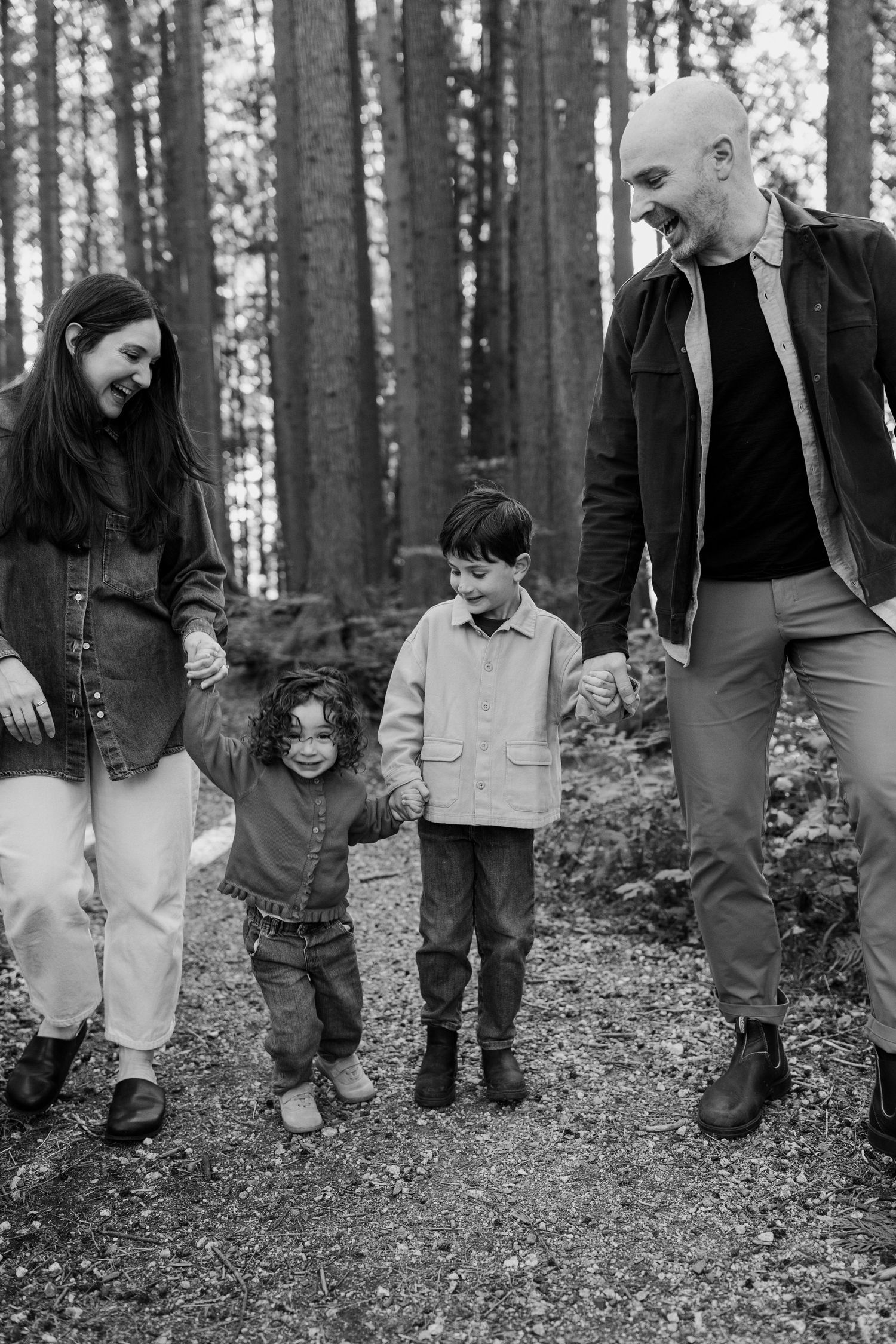 Black and white photo of a happy family of four walking hand-in-hand through a forest trail.