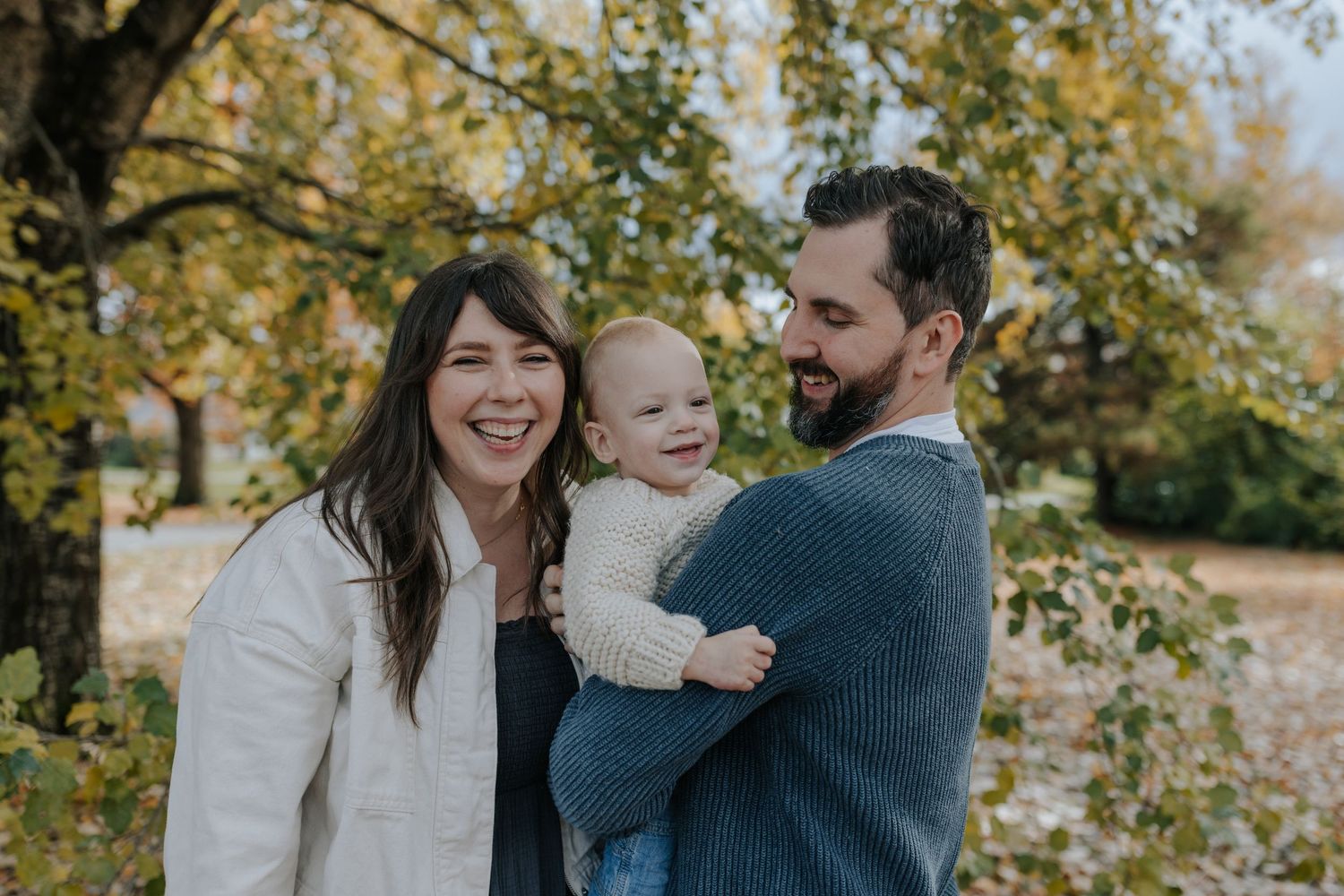 Happy family with baby enjoying autumn park, parents smiling outdoors among golden fall foliage.