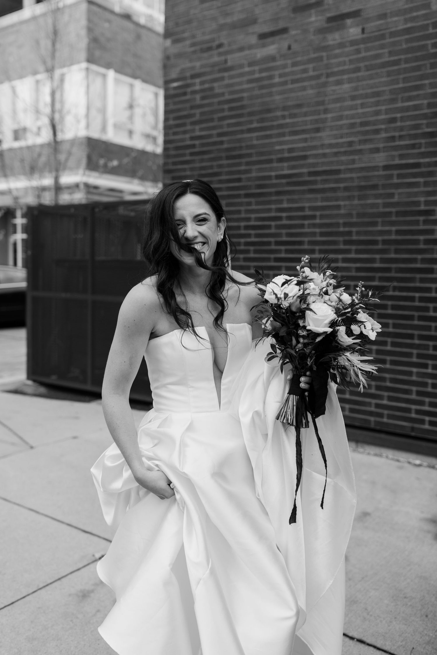A bride in a white wedding dress holds a bouquet while smiling on a city sidewalk.