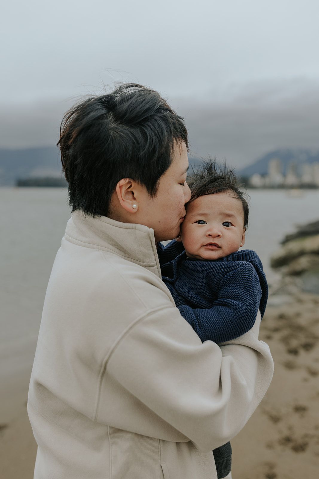 Intimate parent-child moments captured on cloudy beach with mountains in background.