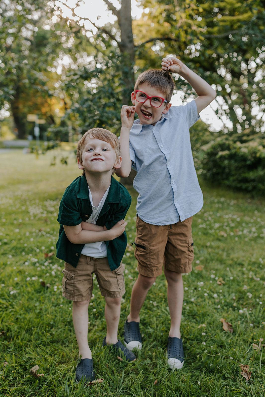 Two children make silly poses and laugh together while playing in a grassy outdoor setting.