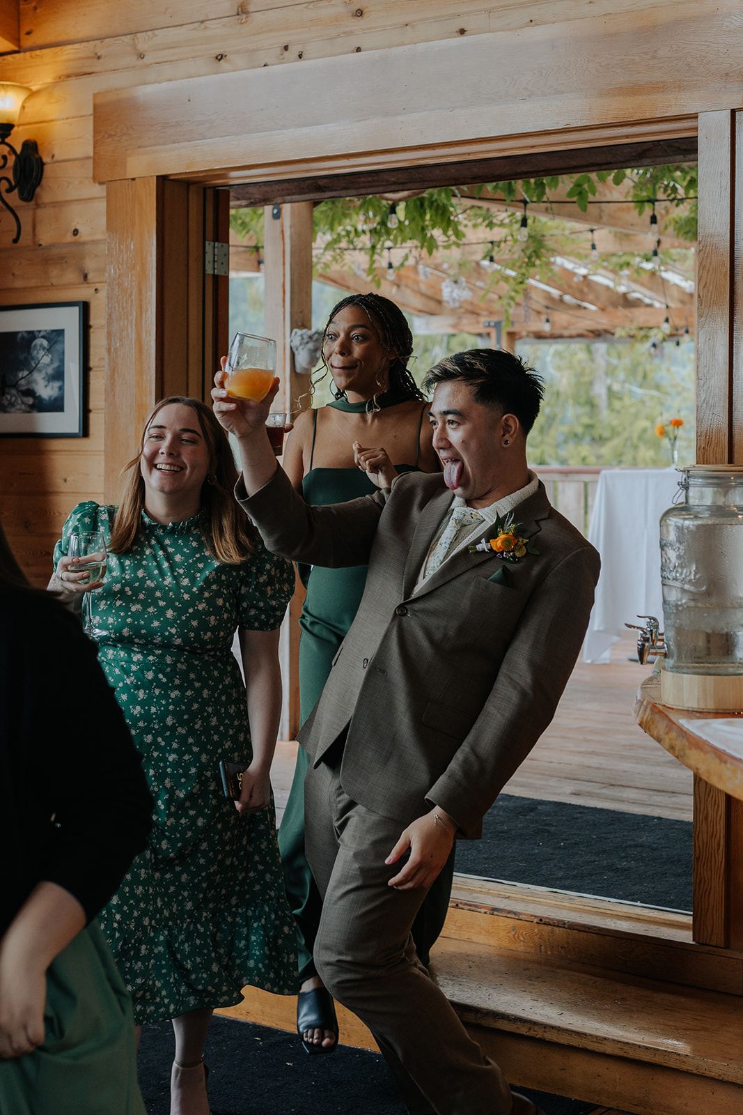 Wedding guests enjoying drinks by large windows in a rustic wooden venue.