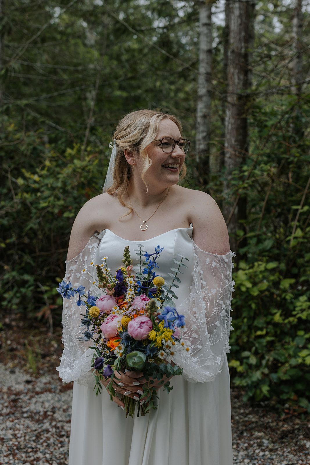 Wedding dress details and floral bouquet featuring pink, blue and yellow blooms in a forest setting.