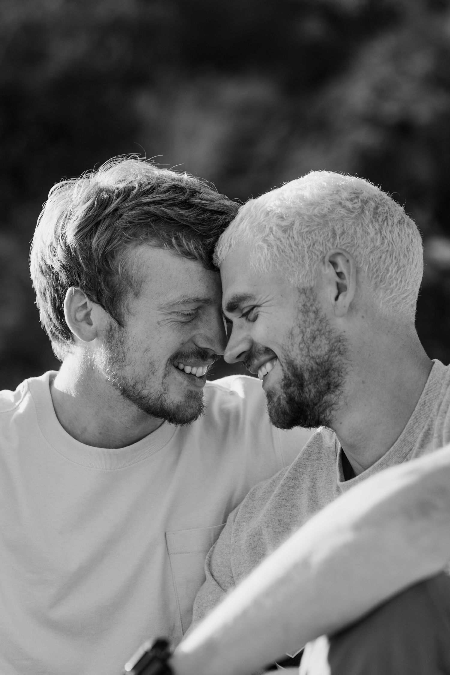 Black and white photo of two smiling men with foreheads touching, sharing a joyful intimate moment outdoors.
