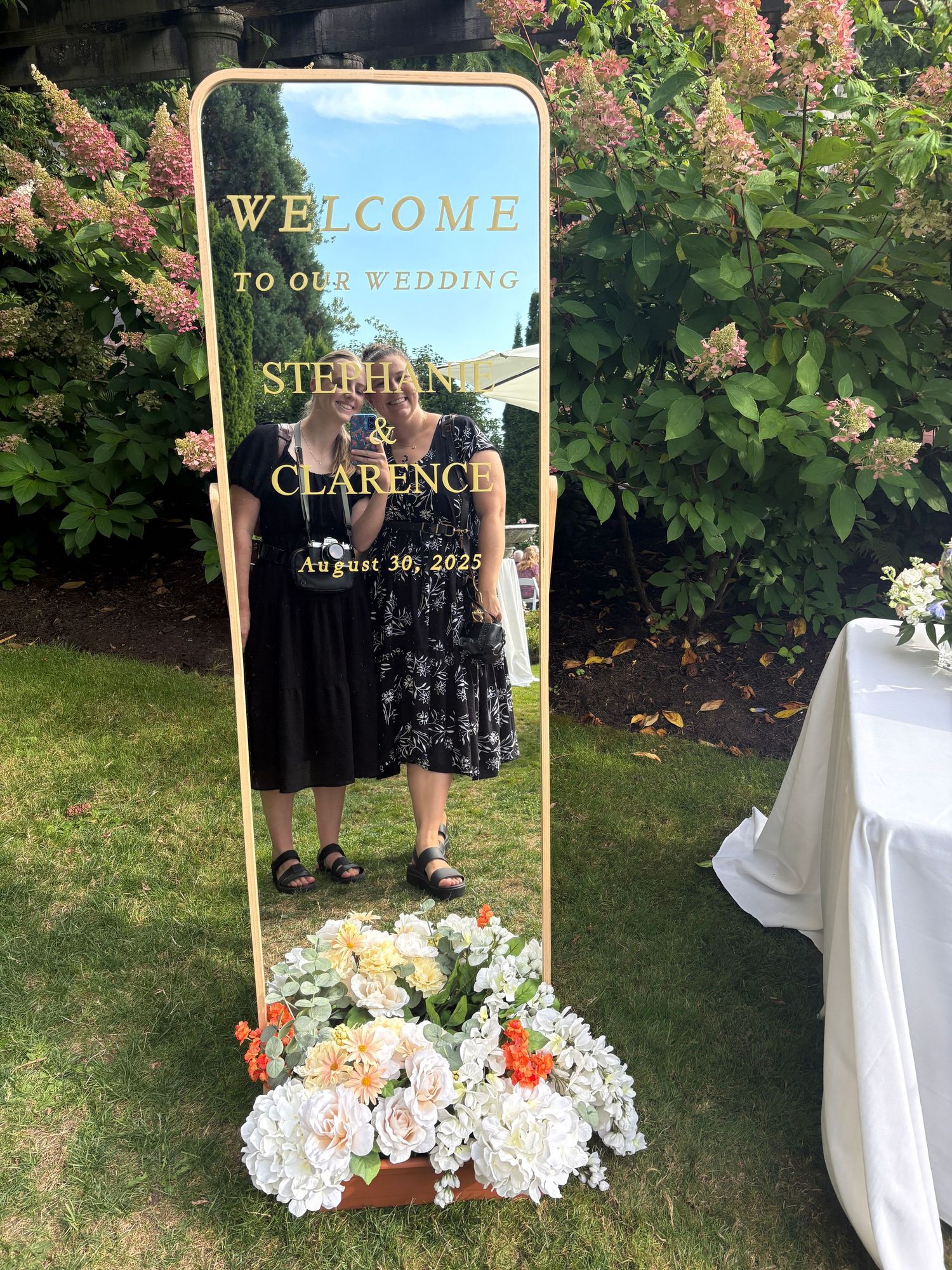 Elegant wedding welcome sign with gold frame and floral decorations on a garden lawn.