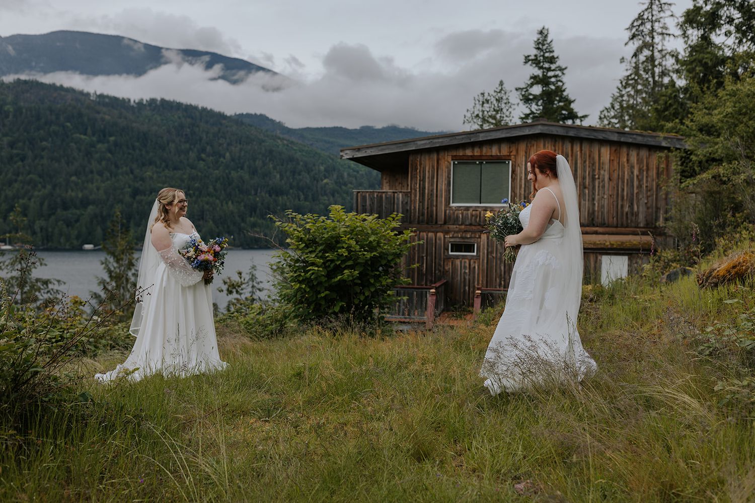 Two brides in white dresses stand in a grassy field with a rustic cabin and mountain lake in the background.